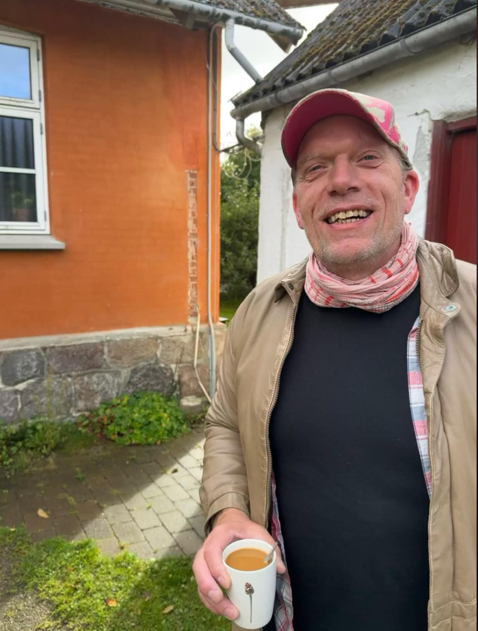 Jens Stardust smiler med en kop kaffe foran et orange hus