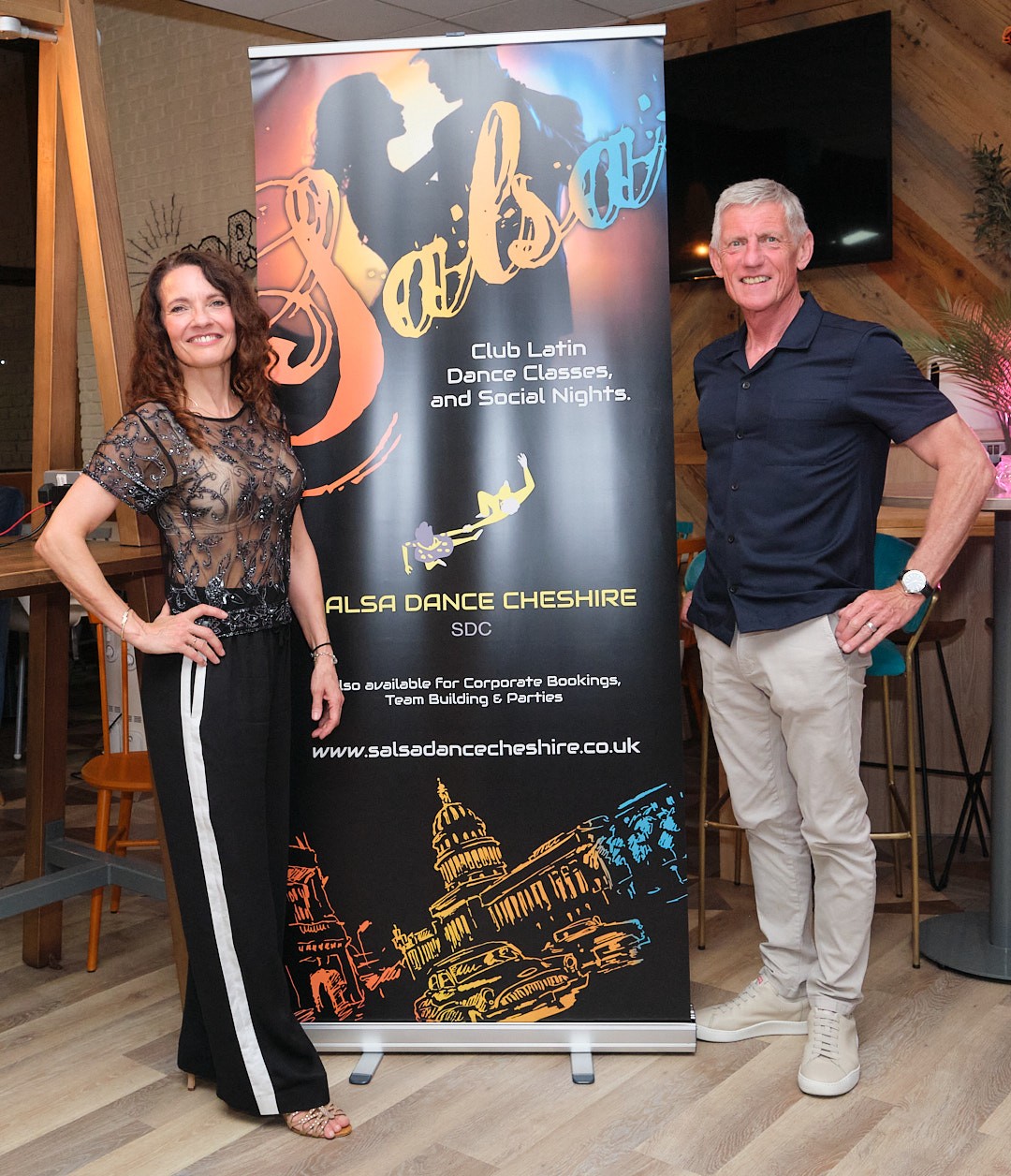 Two smiling salsa teachers standing beside a Salsa Dance Cheshire banner
