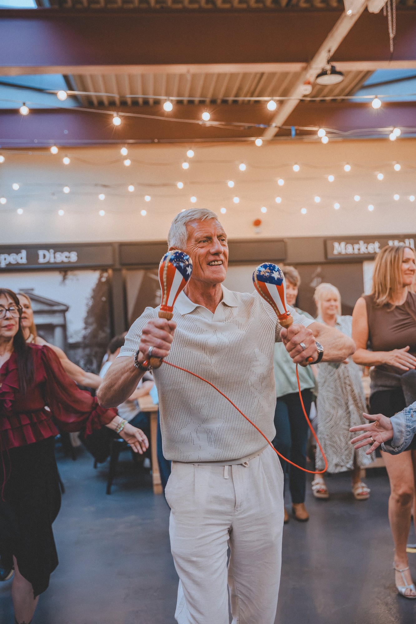 A smiling dancer shaking a pair of maracas under warm string lights at a salsa social