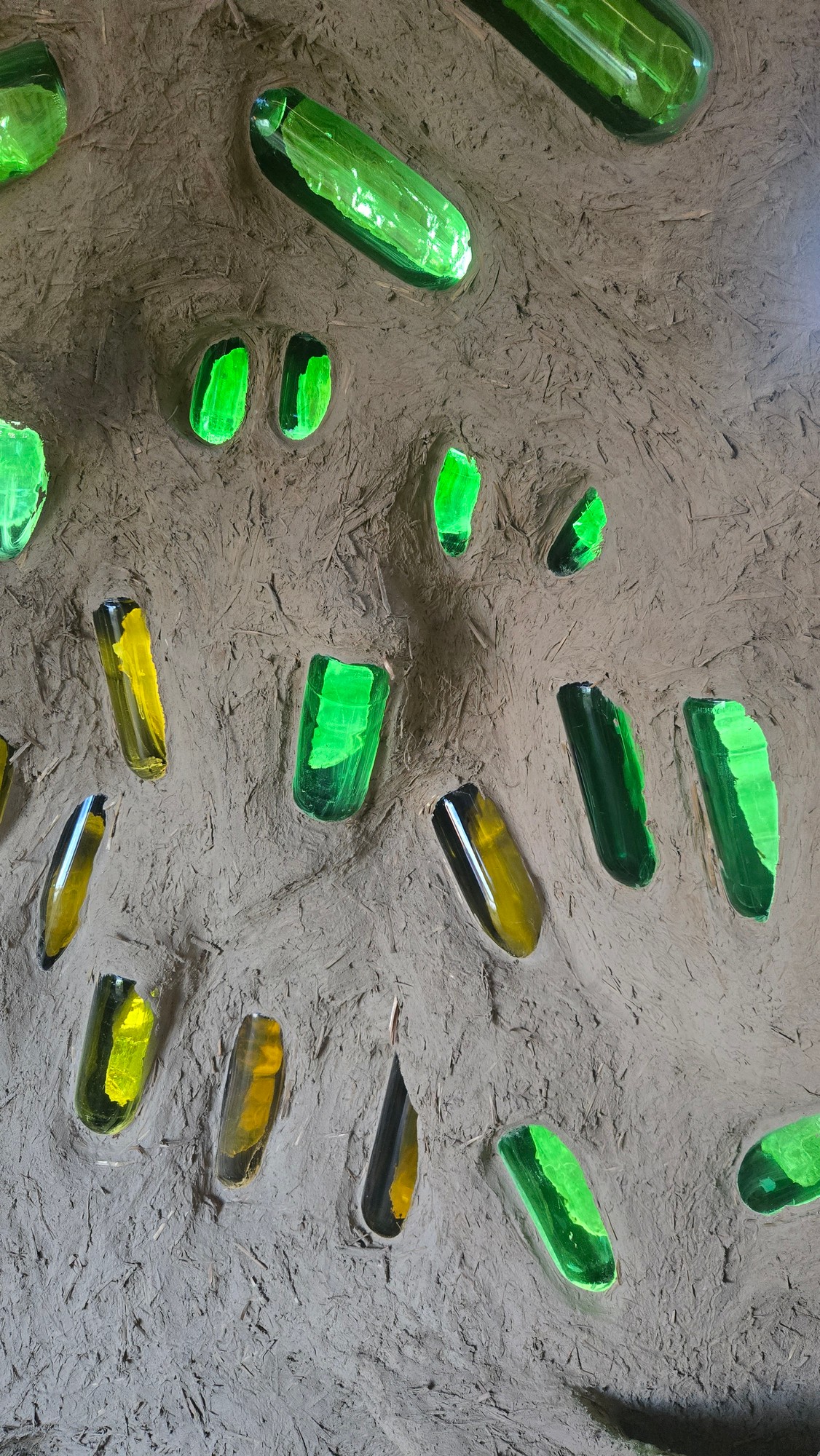 Innenwand der Lehmhütte mit eingelassenen grünen und goldenen Glasflaschen, die Licht durchscheinen lassen