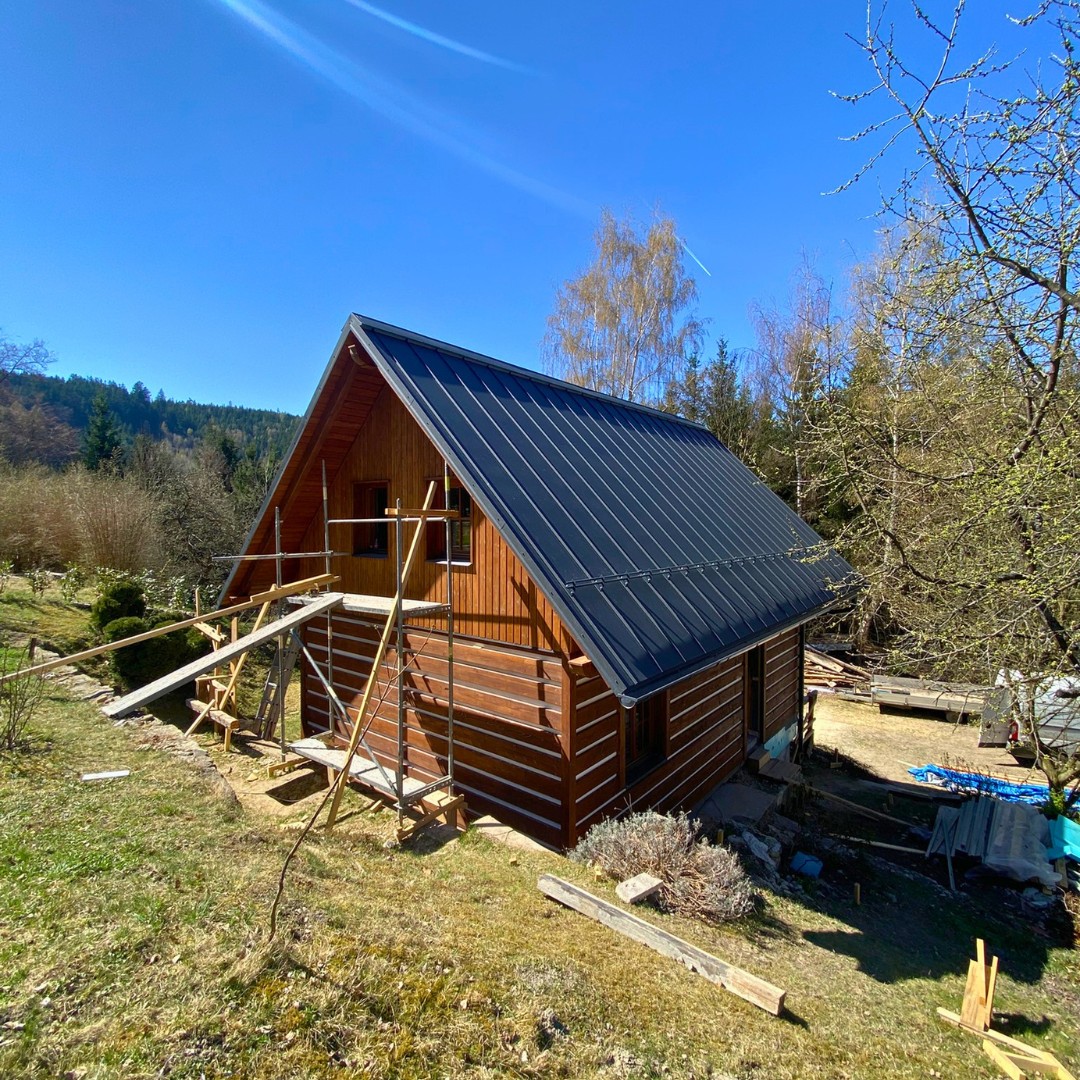 Dokončovaná Česká chata — pohled z boku na dřevěnou fasádu z KVH profilů, tmavou plechovou střechu a lešení při finální úpravě