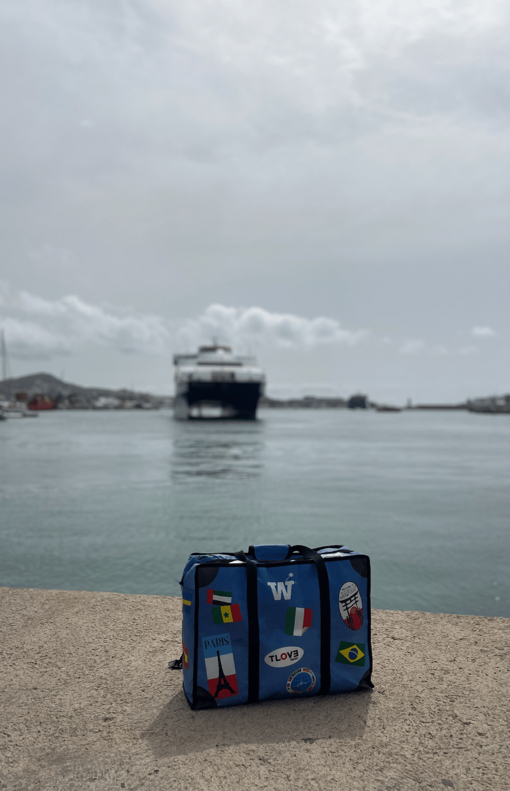 A blue WISSHH Travel Bag with travel stickers on the harbor pier as the ferry arrives in the background