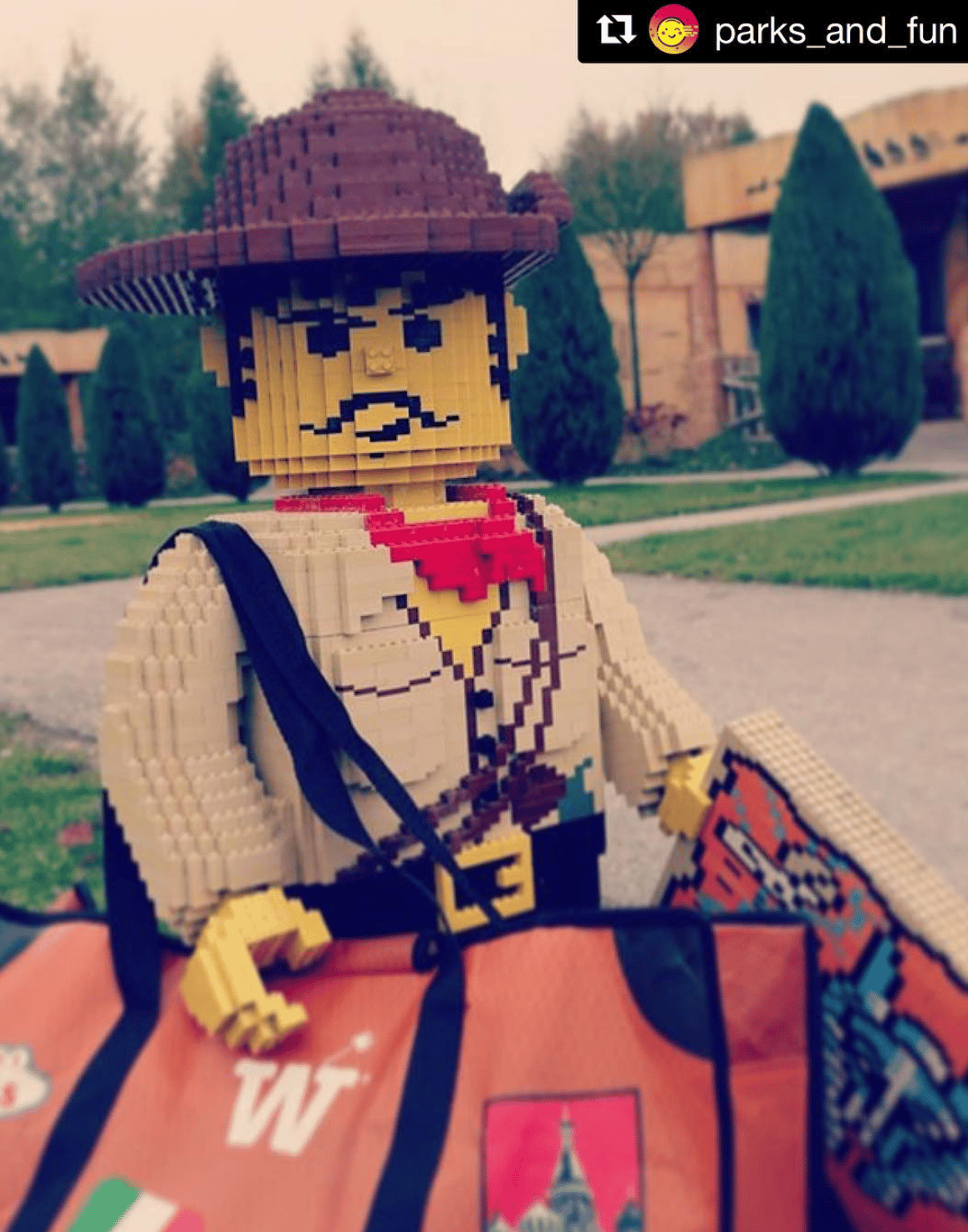 A giant LEGO explorer figure carrying the orange WISSHH Travel Bag with travel stickers at an amusement park