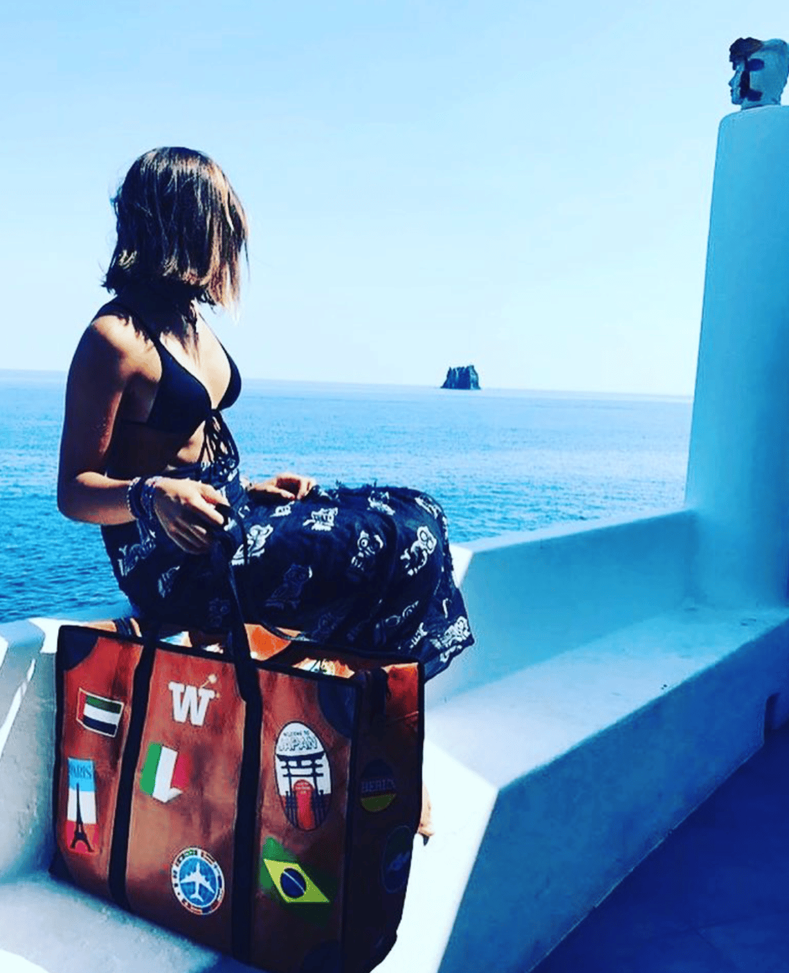 A woman sitting on a white seaside terrace looking at the sea, with the orange WISSHH Travel Bag with travel stickers beside her
