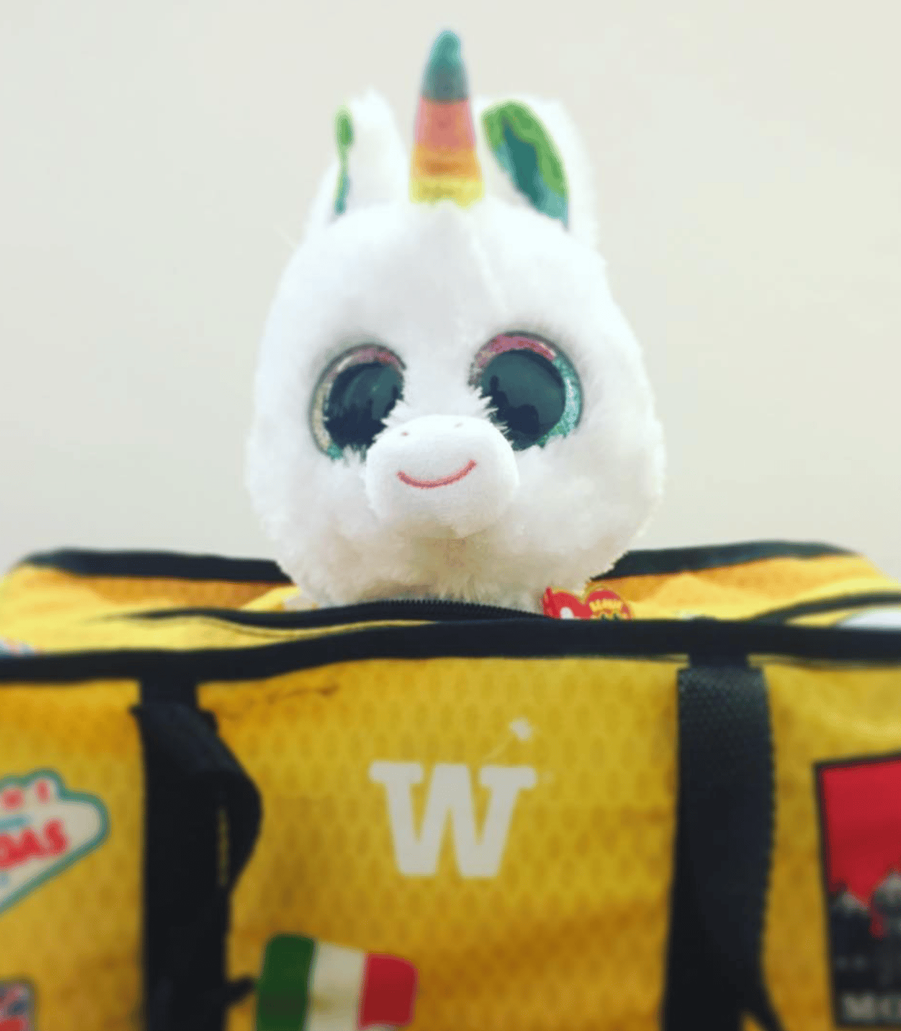 A plush unicorn peeking out of a yellow WISSHH Travel Bag with travel stickers