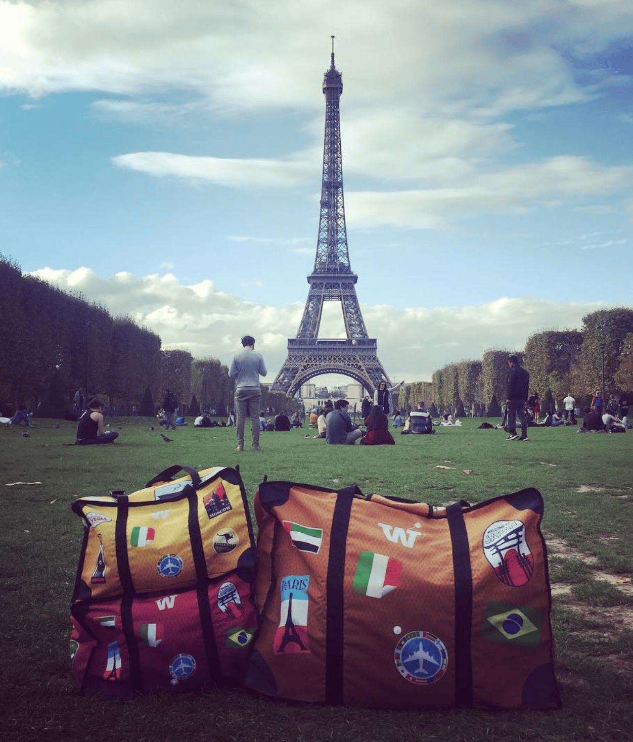 Colorful yellow, red and orange WISSHH Travel Bags with travel stickers on the lawn in front of the Eiffel Tower in Paris