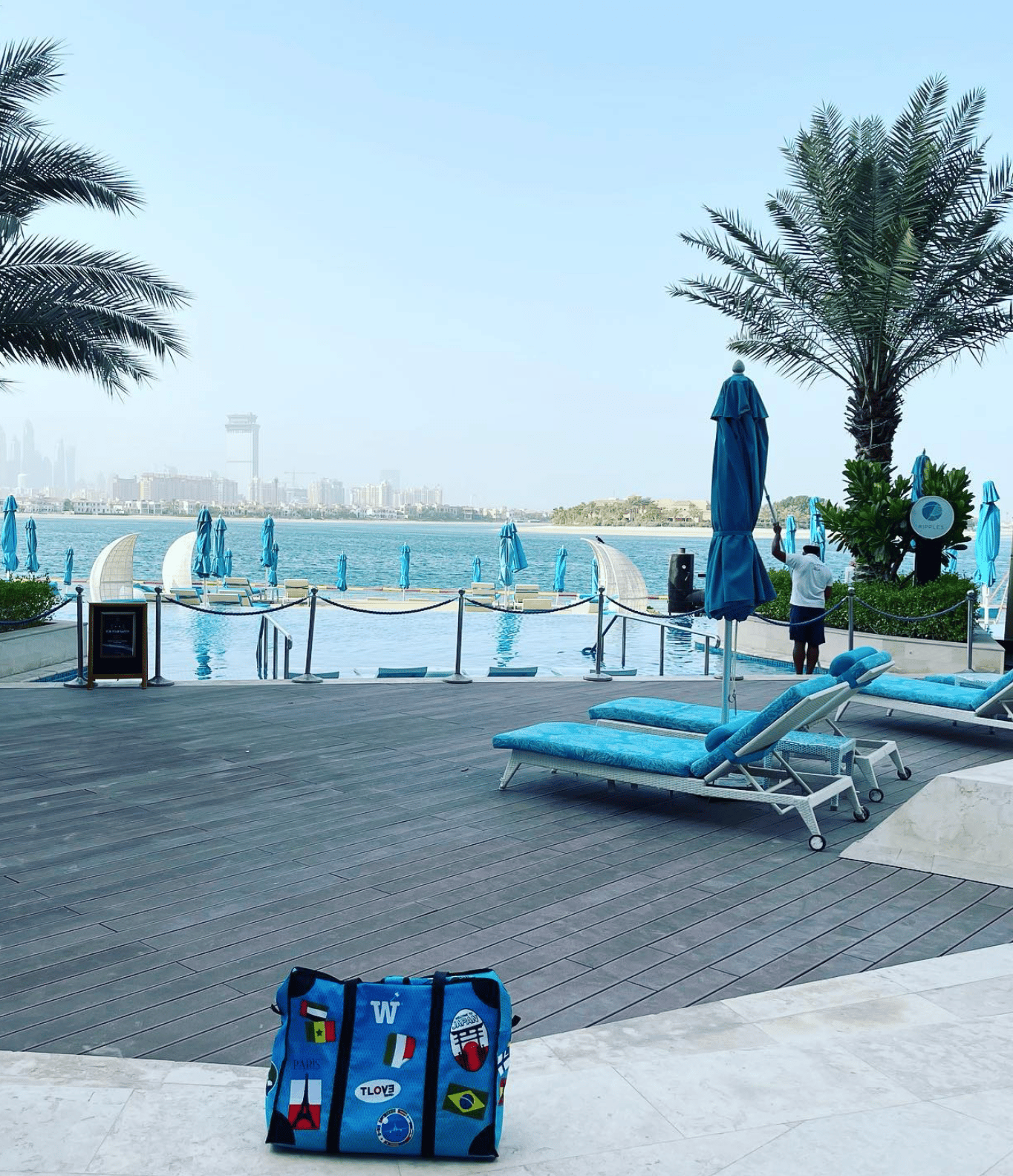 A blue WISSHH Travel Bag with travel stickers by the pool of a beach club with the Dubai skyline behind