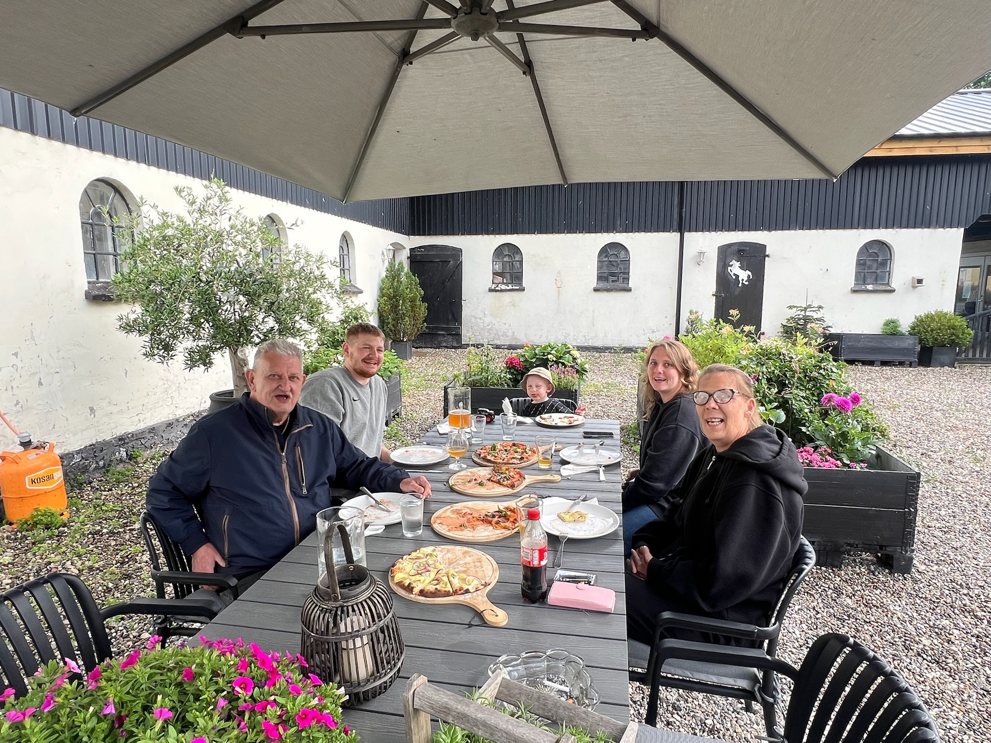 Selskab spiser pizza ved det lange bord under parasollen på gårdspladsen