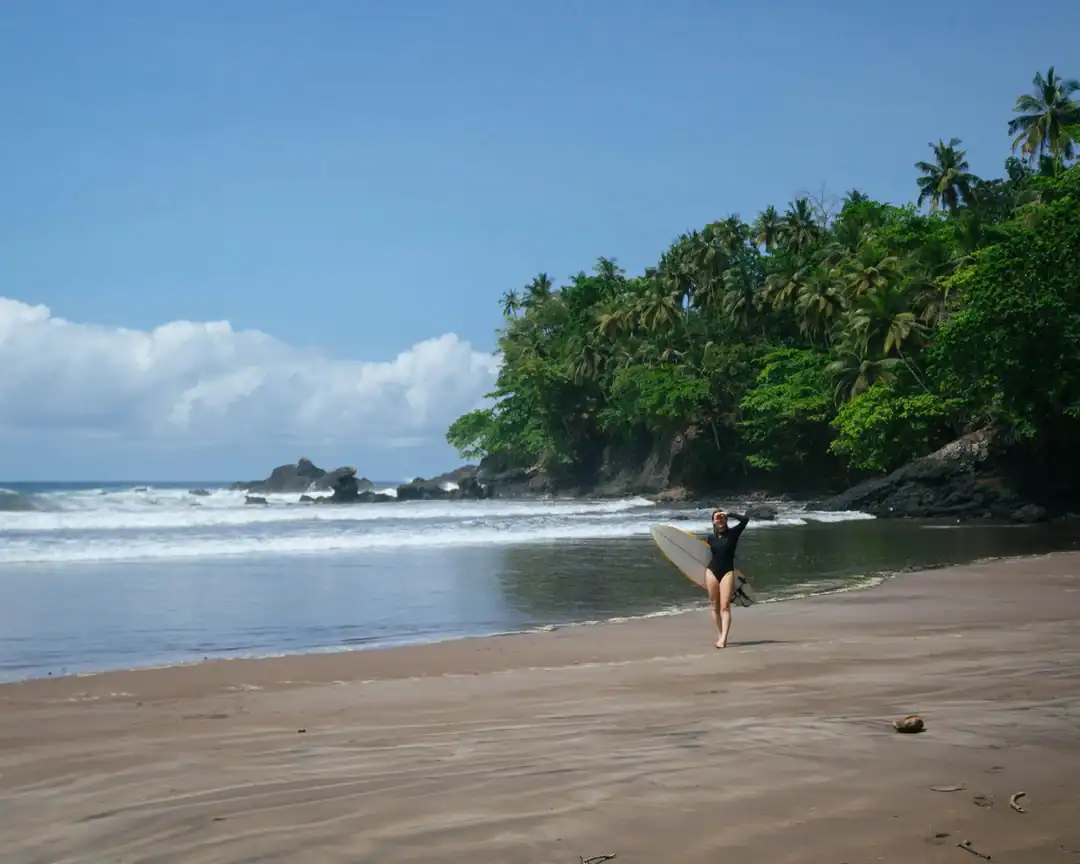 Surfista a carregar uma prancha na praia de areia escura de Sete Ondas em São Tomé, com ondas e floresta atrás