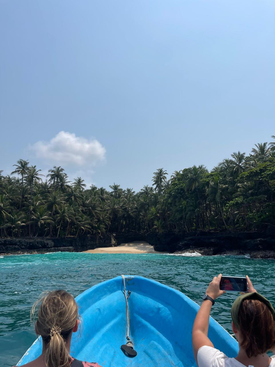 Vista da proa de um barco azul a aproximar-se de uma enseada de praia isolada ladeada por palmeiras altas e rocha vulcânica negra