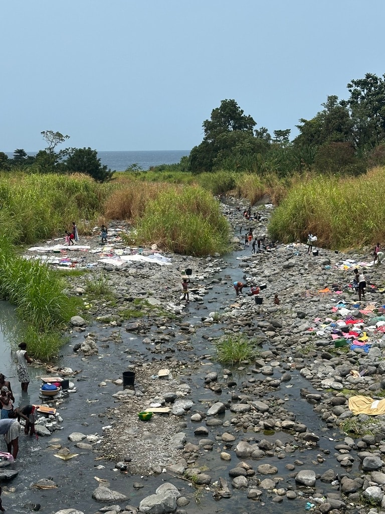 Famílias a lavar roupa num rio rochoso largo na estrada sul de São Tomé, com o oceano visível ao fundo