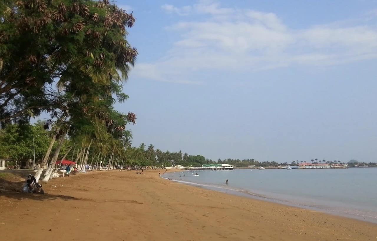 Praia Emília perto da cidade de São Tomé, uma praia com palmeiras onde os locais nadam e um cais é visível ao fundo