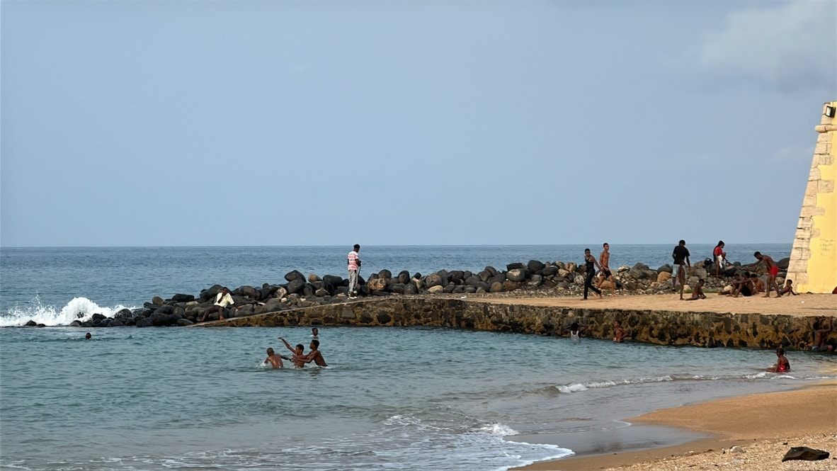 Crianças a nadar na Praia PM na cidade de São Tomé, com a muralha do antigo forte e a água azul calma