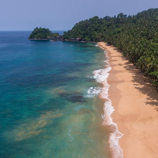 Vista aérea da Praia Jalé na costa sul de São Tomé, mostrando areia dourada, água turquesa e promontório coberto de palmeiras