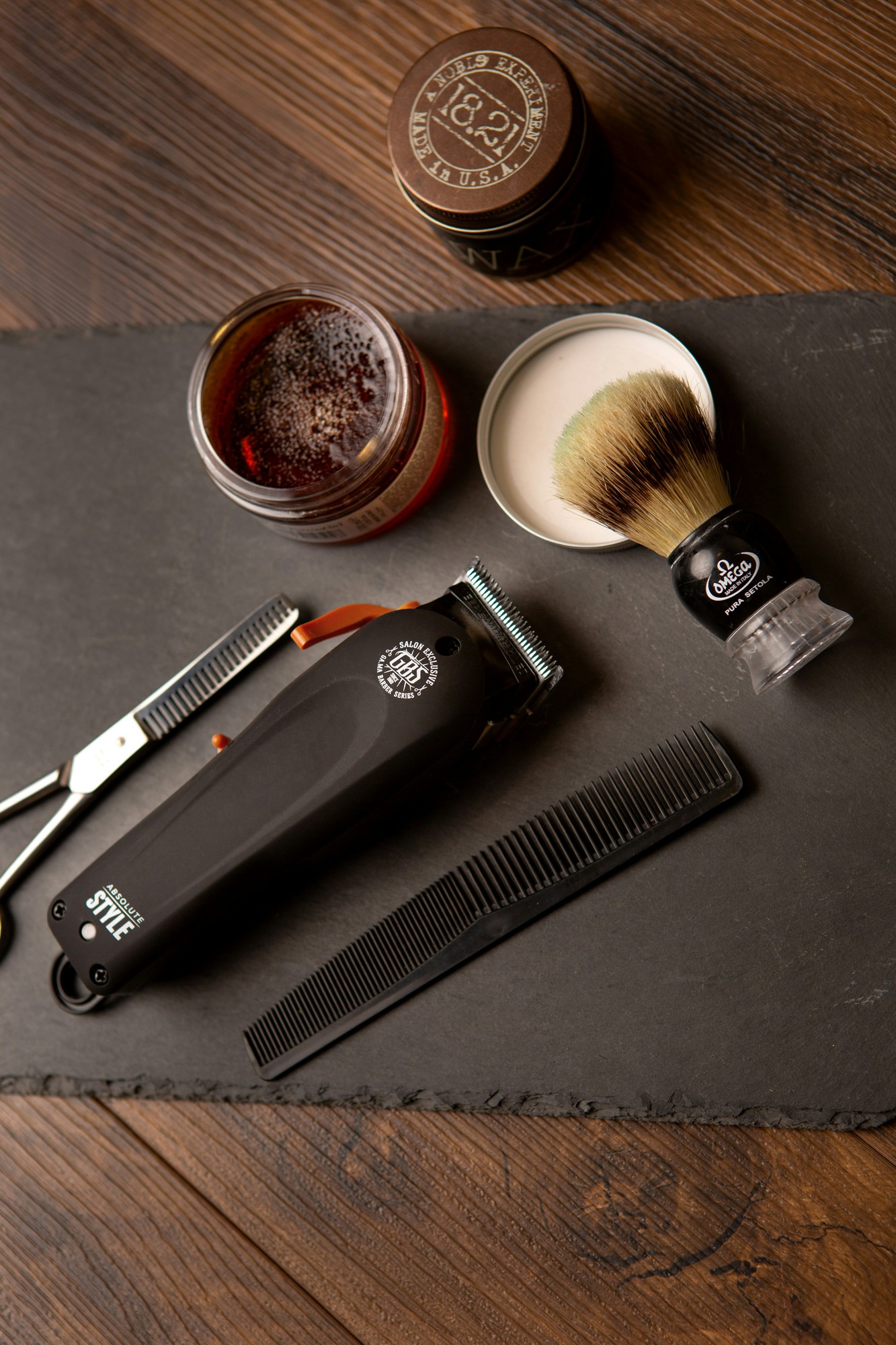 Clipper, shears, comb, pomade and shaving brush laid out on slate