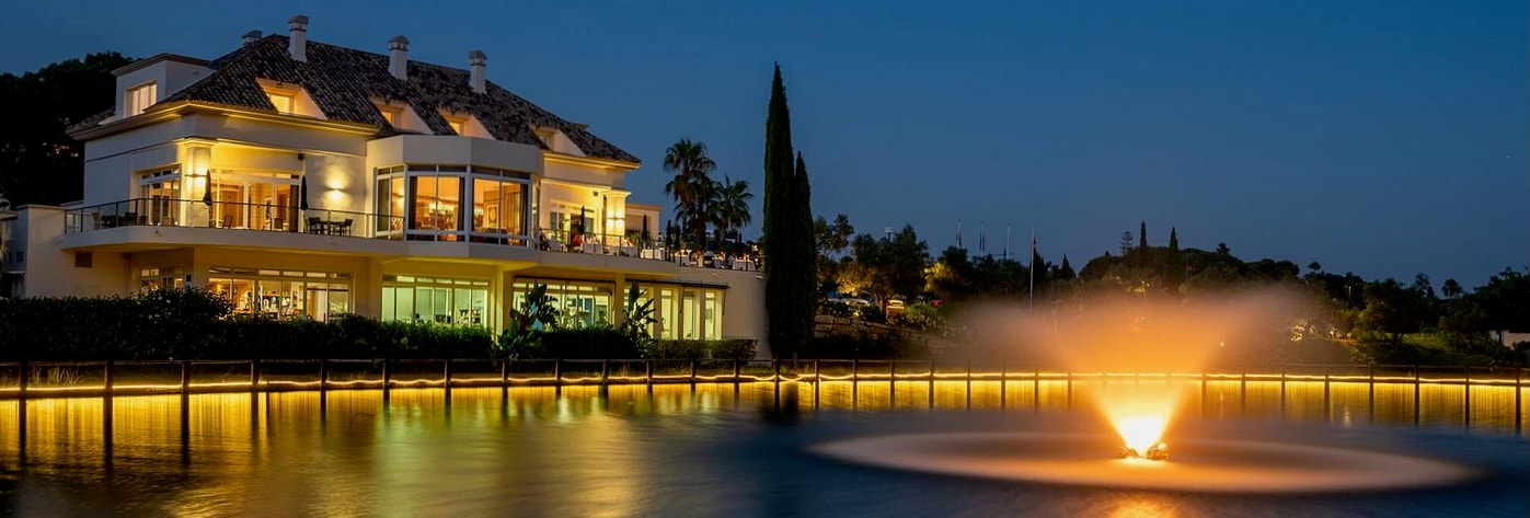 El Lago at twilight beside the lake, with its windows glowing and the fountain illuminated across the water