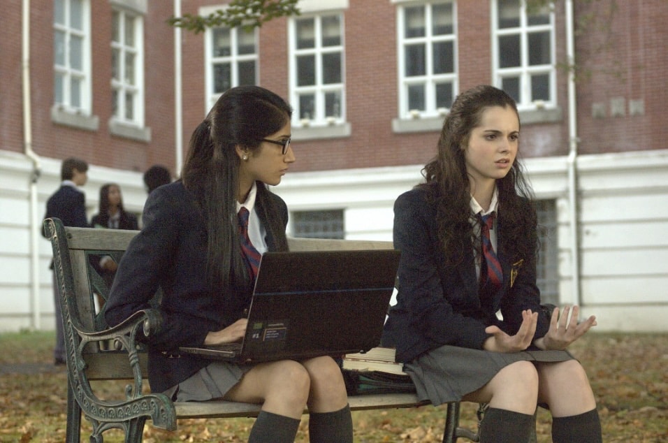 Two students on a bench with a laptop outside a brick school building in Restless Virgins