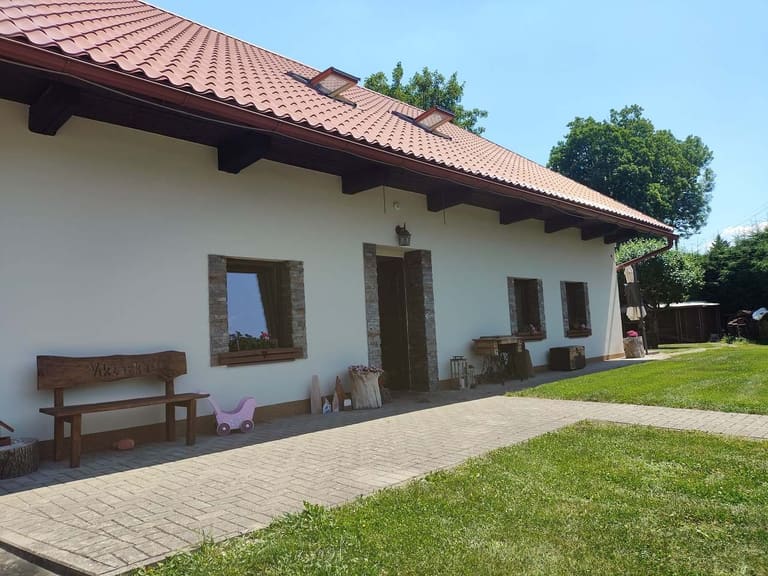 Biała fasada domku z kamiennymi opaskami okiennymi i drzwiami wejściowymi, przed nimi drewniana ławka z napisem Viktorka