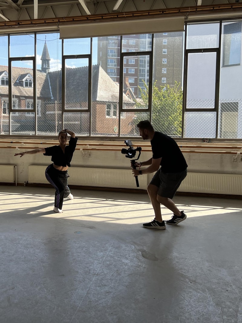 A camera operator films a dancer mid-movement in a sunlit dance studio