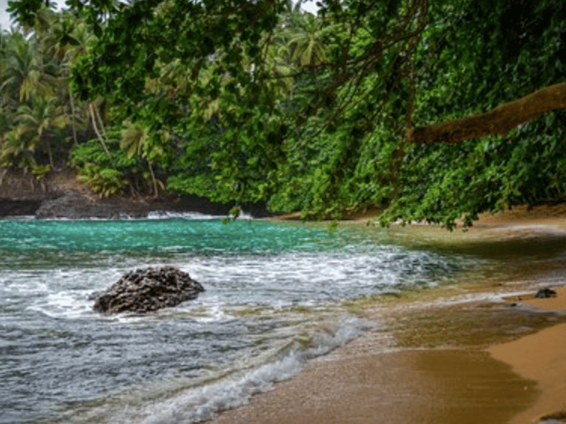 A enseada abrigada da Praia Piscina no sul de São Tomé, com água turquesa, uma rocha vulcânica e floresta densa a enquadrar a praia