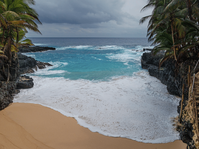 Enseada da Praia Bateria em São Tomé, enquadrada por rochas vulcânicas negras e palmeiras com água turquesa