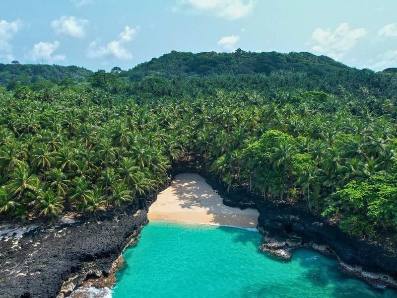 Aerial view of Praia Bateria on São Tomé, showing a secluded cove with turquoise water surrounded by black volcanic rocks and dense palm forest