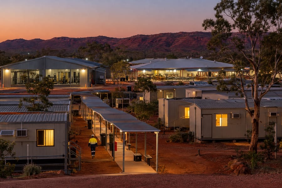 A FIFO mining camp village at dusk with demountable accommodation rooms, mess hall and gym buildings on red dirt in the Australian outback