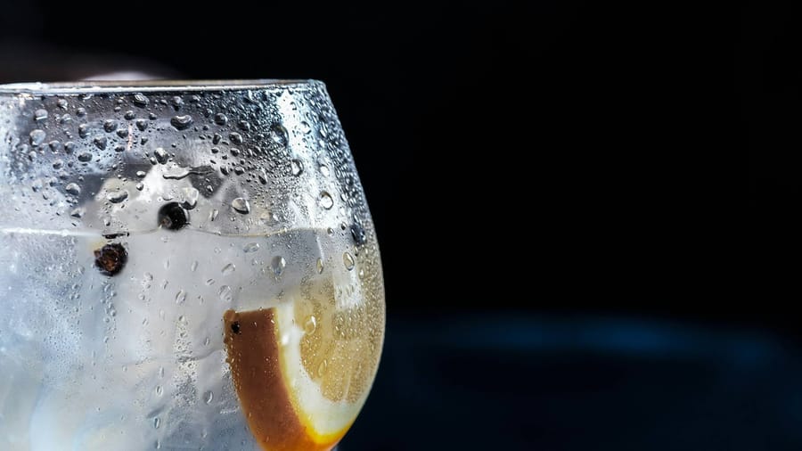 A close-up of a glass of gin and tonic from Cape Byron Distillery, filled with ice cubes, a slice of lemon, and juniper berries, with condensation on the glass.