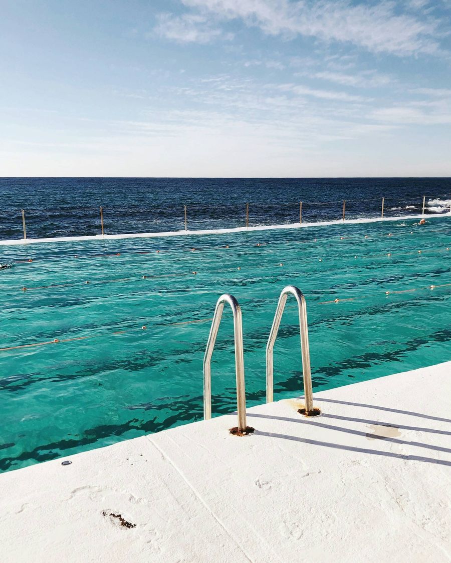 A serene ocean pool with clear turquoise water, bordered by a white concrete deck and metal pool ladder. The ocean and horizon extend into the background under a lightly clouded blue sky.