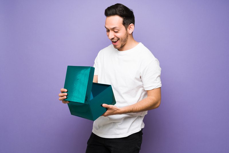 Excited and thrilled man with a beaming smile opening a birthday present while looking surprised and shocked. Latin man holding a gift box