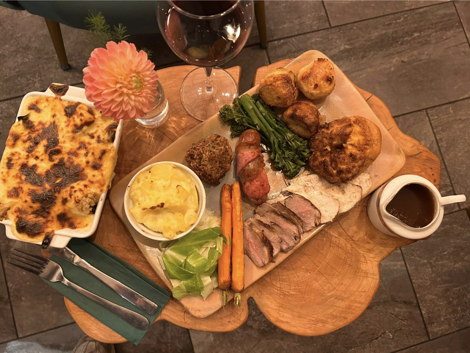 A rustic wooden board with roast dinner, cauliflower cheese, carrots and a glass of red wine