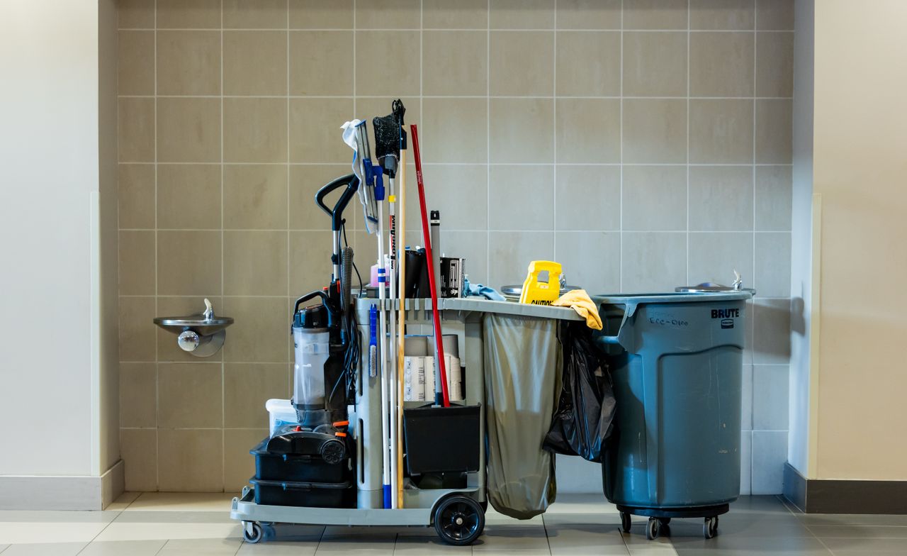 Janitorial cleaning trolley stocked with mops, vacuum, bins and supplies beside a tiled wall
