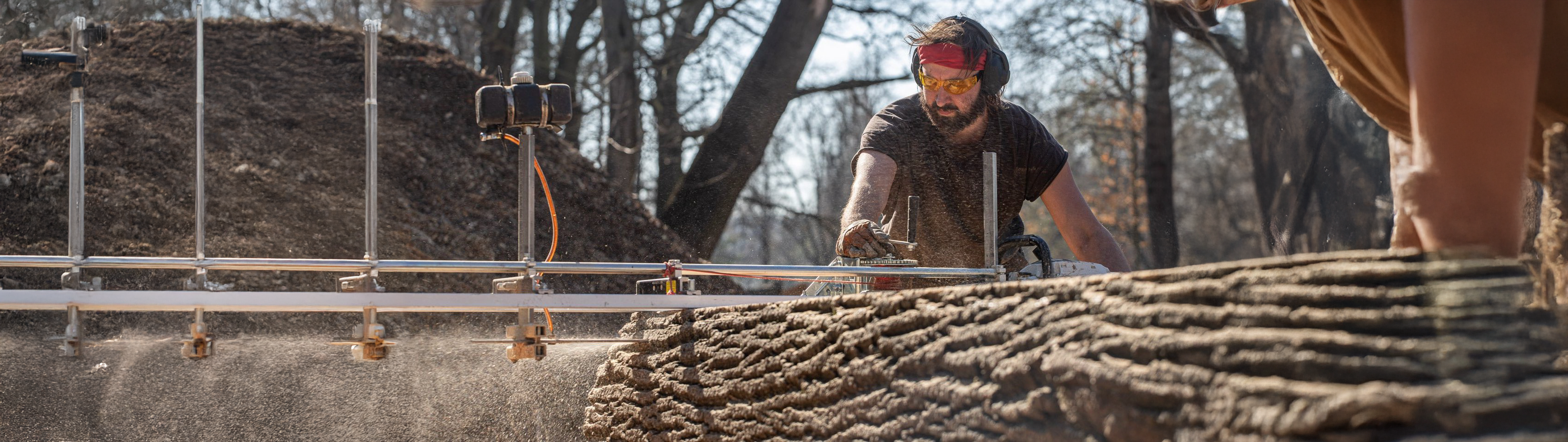 Řemeslník v ochranných brýlích precizně řeže kmen mobilní pilou v přírodním prostředí