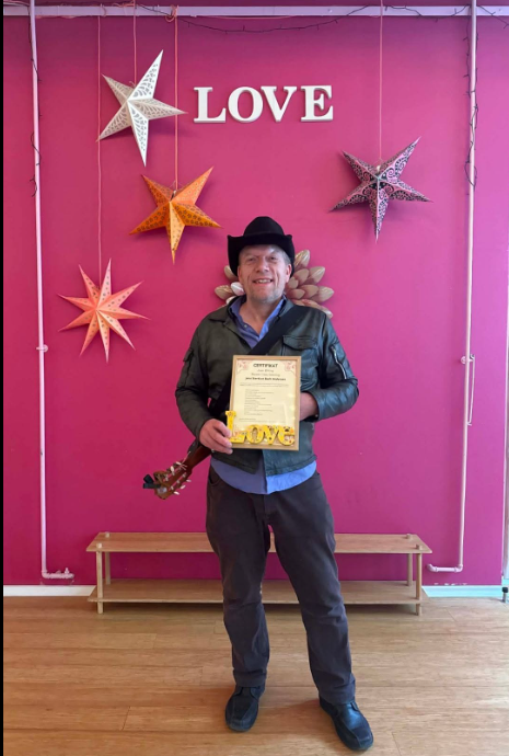 Jens Stardust smiler med hat og guitar og viser et bevis foran en lyserød væg med LOVE-skilt