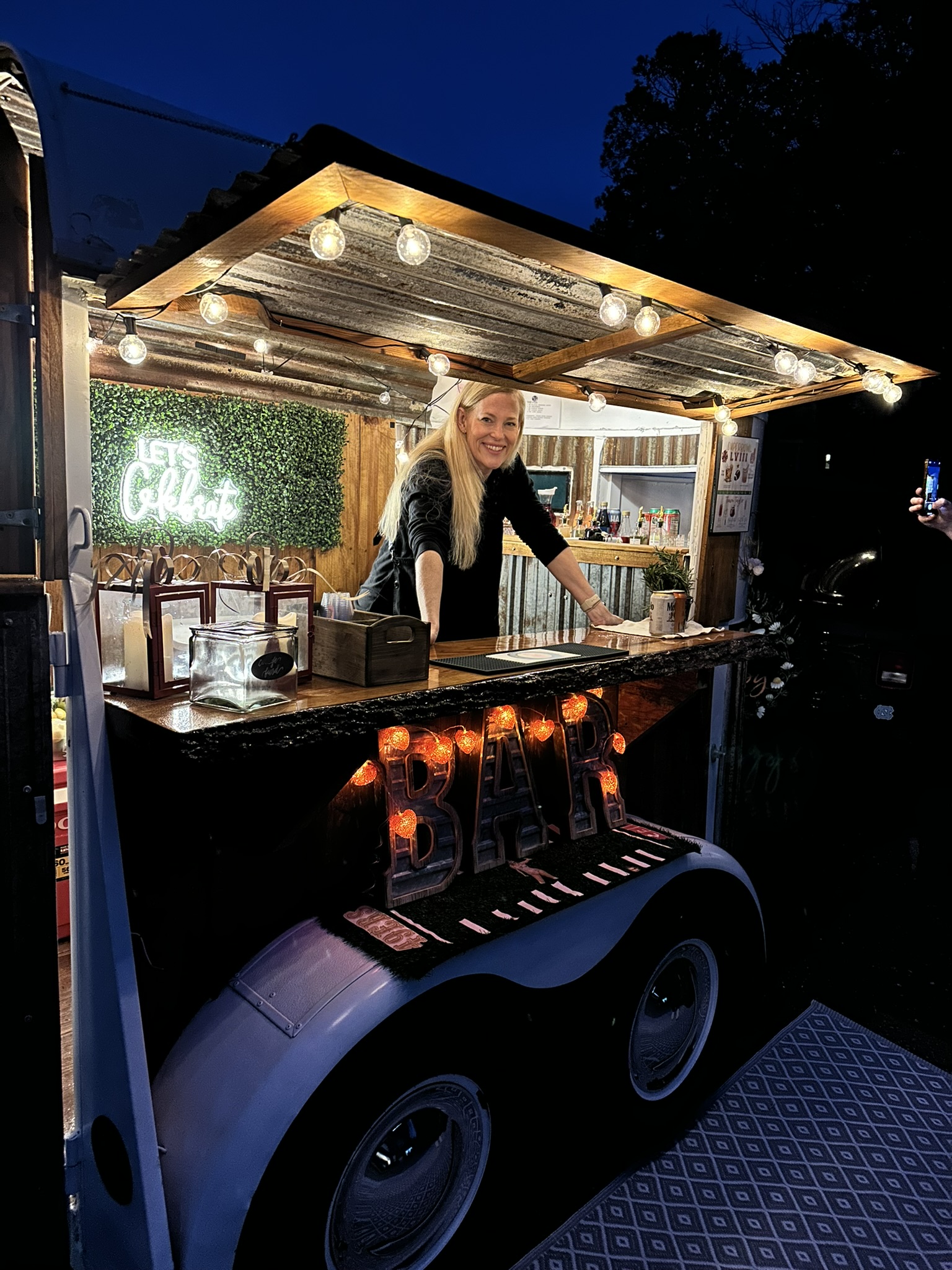 Lisa smiling behind The Hitching Post mobile bar trailer at dusk with light-up BAR letters and string lights