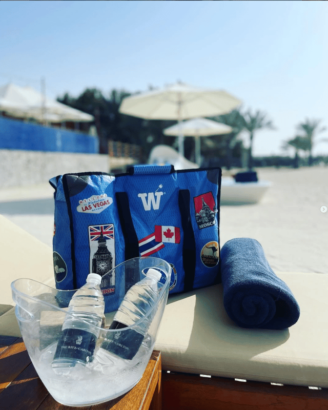 A blue WISSHH Travel Bag with stickers beside water bottles and a rolled towel at a beach club