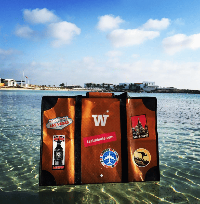 An orange WISSHH Travel Bag in shallow transparent water near a beach club