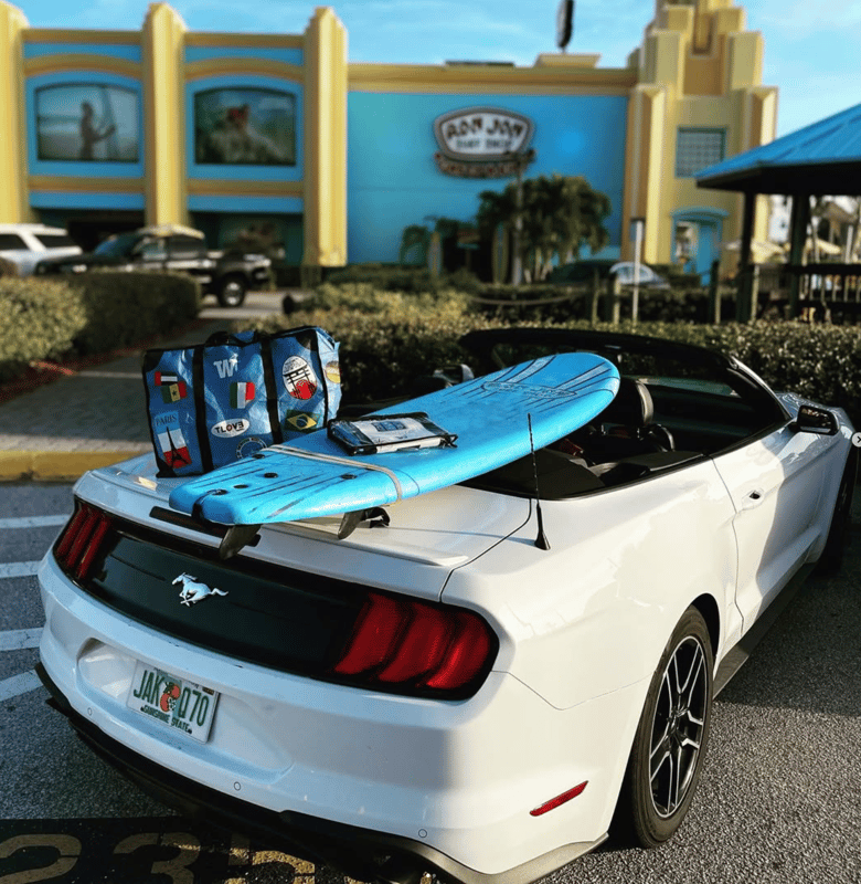 A white convertible with a surfboard and WISSHH bags on the back seat, parked in front of a surf bar