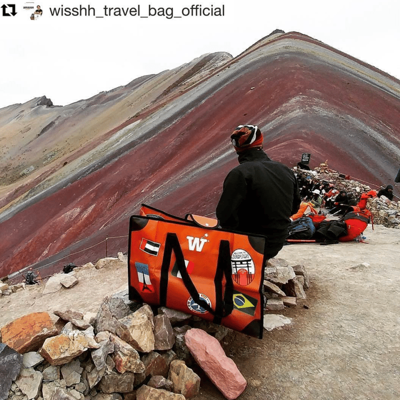 An orange WISSHH Travel Bag with stickers resting on rocks at Rainbow Mountain, Peru