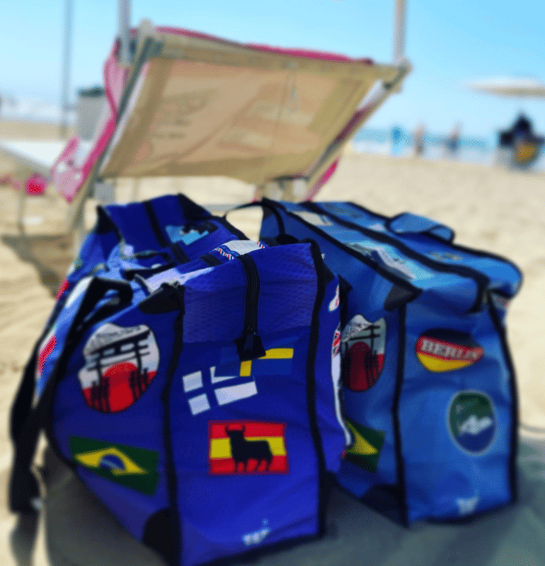 Blue WISSHH duffels with world flags lined up on the sand at the beach