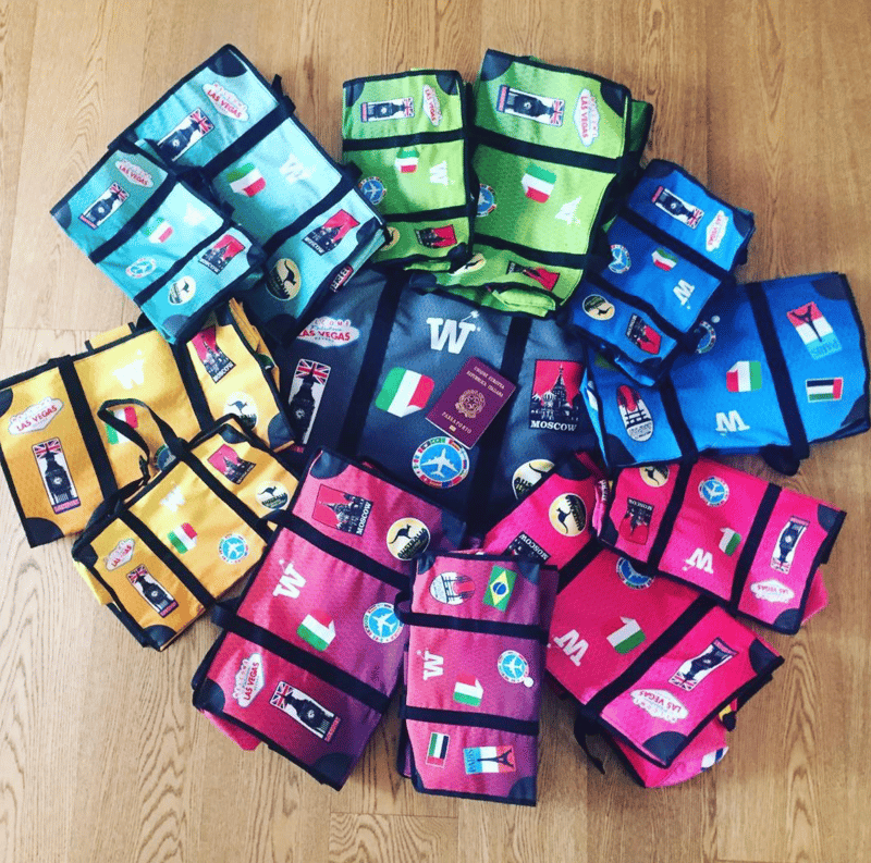 Many colorful WISSHH Travel Bags with stickers arranged in a circle on a wooden floor