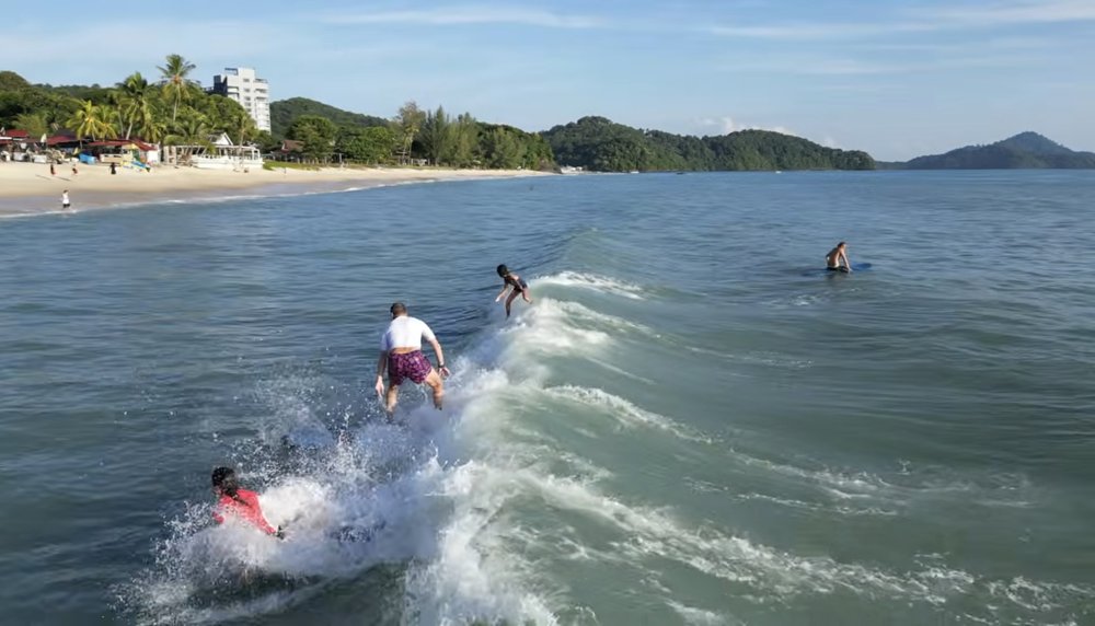 Several surfers, including two children, riding and swimming through a wave together at Pantai Tengah, Langkawi, with the beach and a resort visible in the background