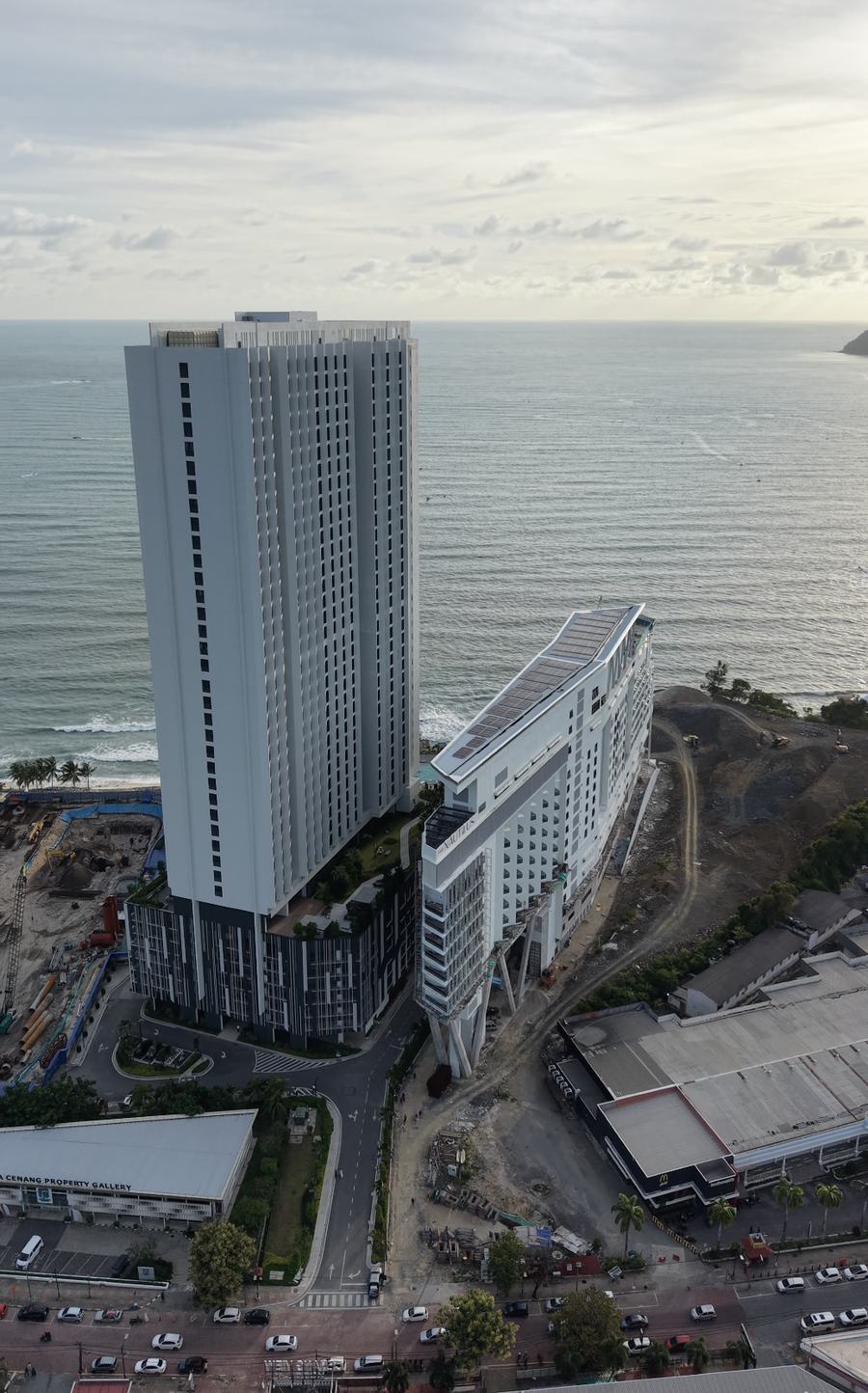 A tiered white building under a setting sun, its upper floors still wrapped in scaffolding, standing beside Tropicana's taller completed tower