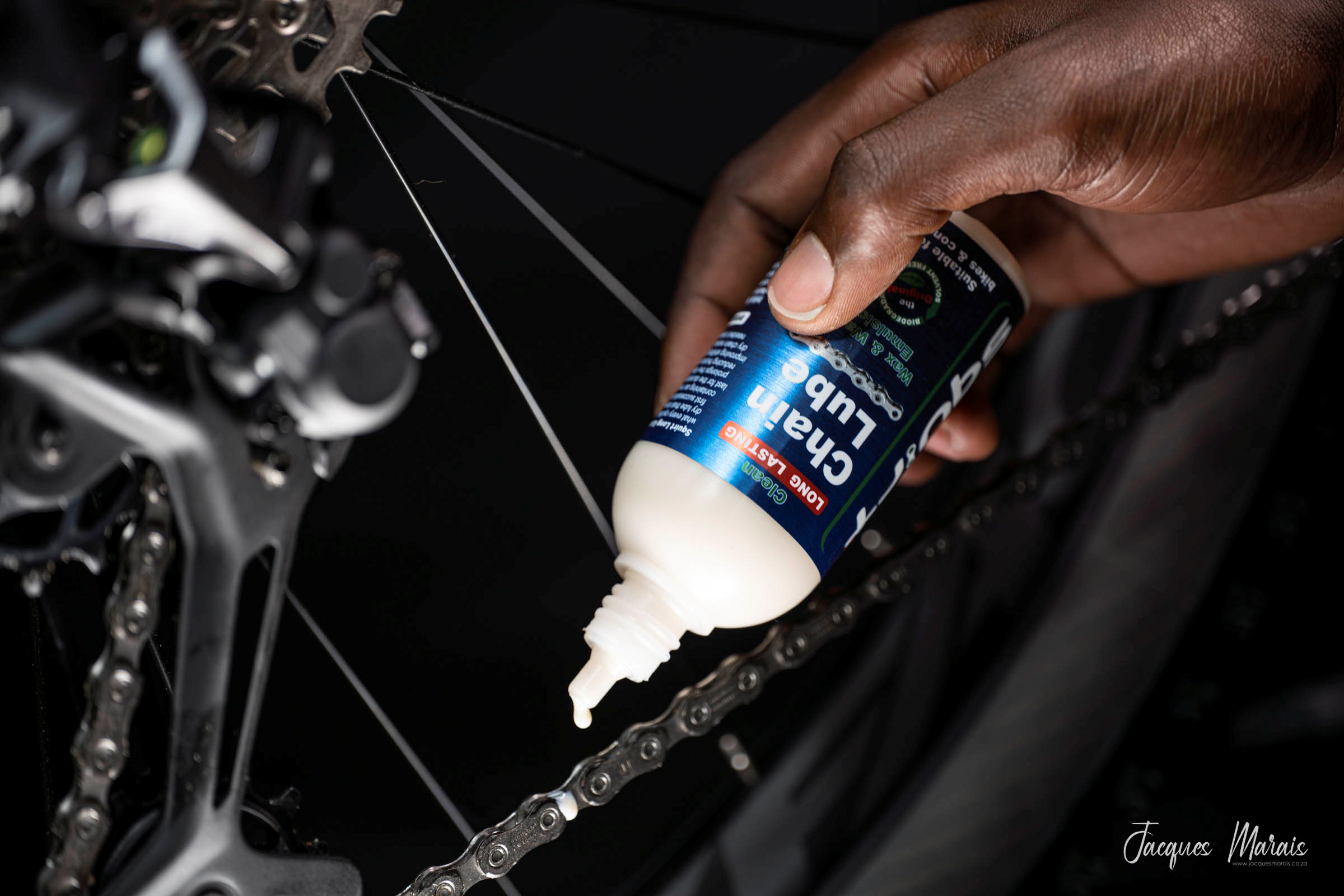 Hånd påfører Squirt Chain Lube på en cykelkæde med mørk carbonramme i baggrunden